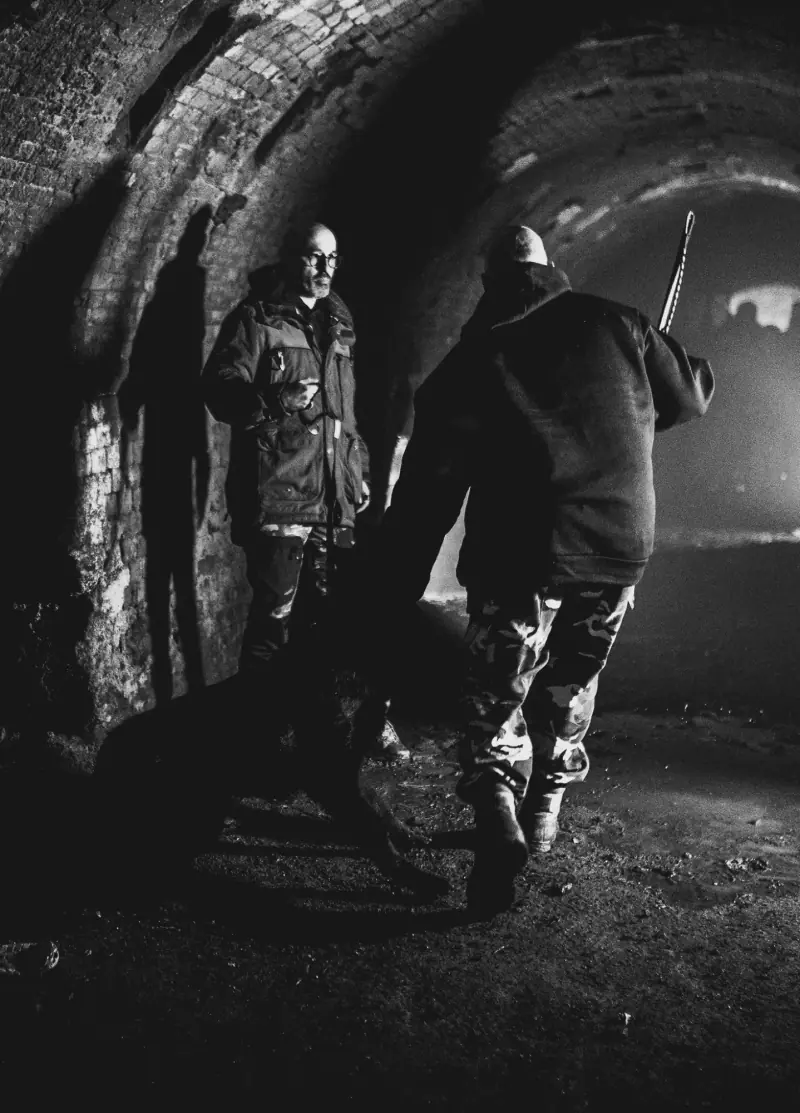 Handler and dog facing a person in protective clothing in a brick passageway.